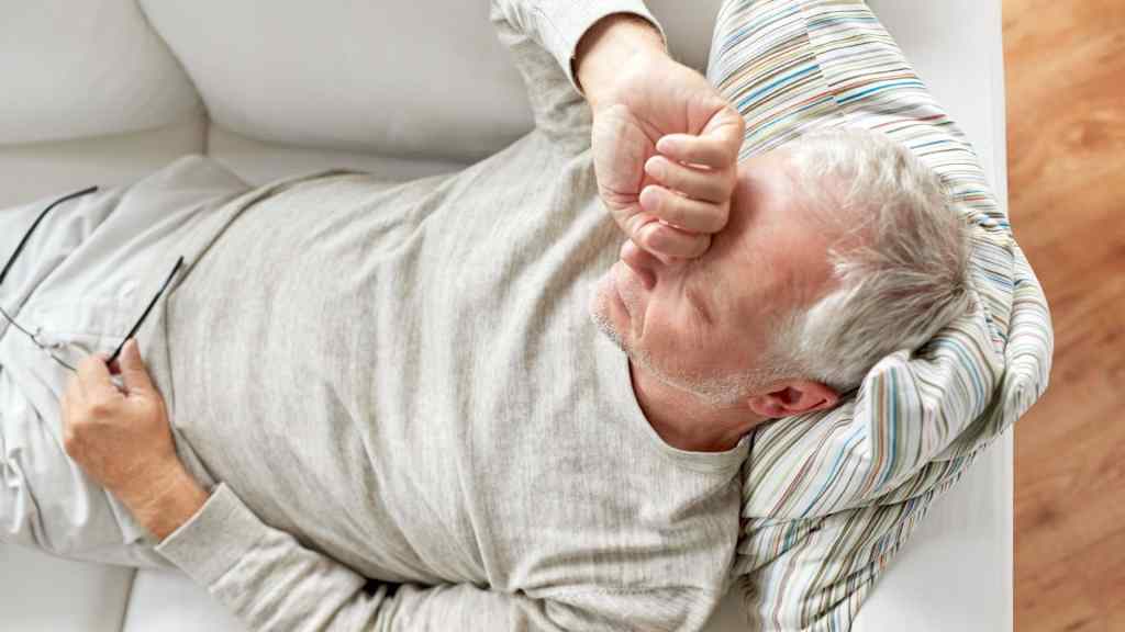 An older man with gray hair lies on a couch with his arm covering his eyes and holds eyeglasses in one hand, appearing tired or unwell.