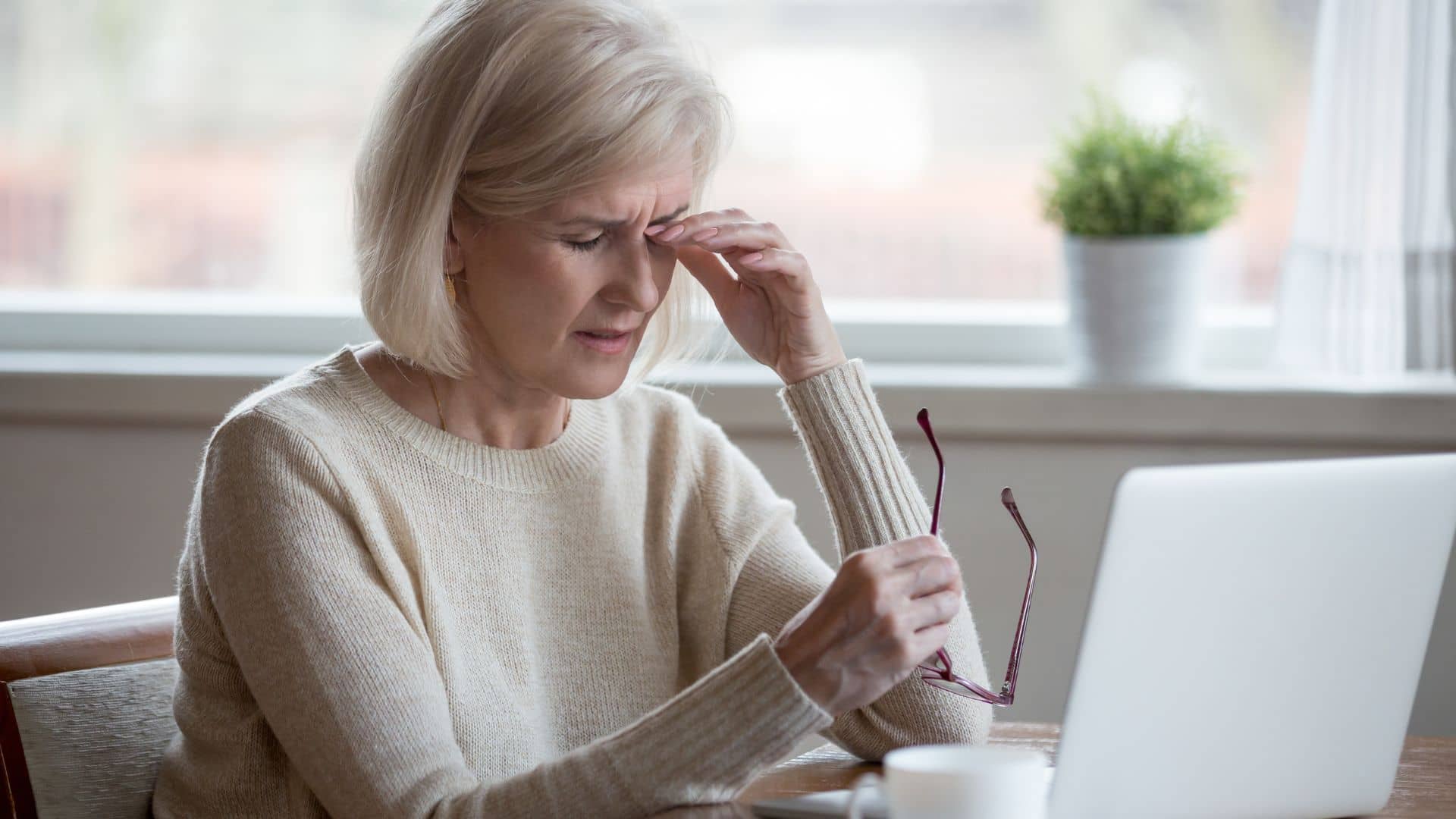 An older woman sitting at a table looks stressed, holding her glasses in one hand and touching her forehead with the other, near a laptop and a potted plant.
