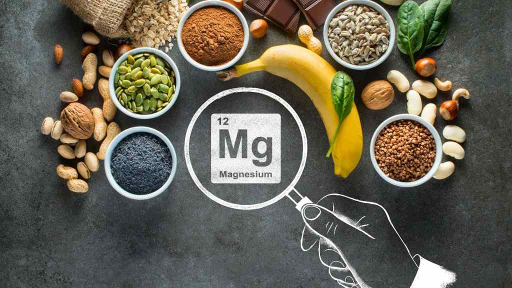 Various magnesium-rich foods such as seeds, nuts, banana, grains, and leafy greens are arranged on a dark surface with a magnifying glass illustration highlighting magnesium.