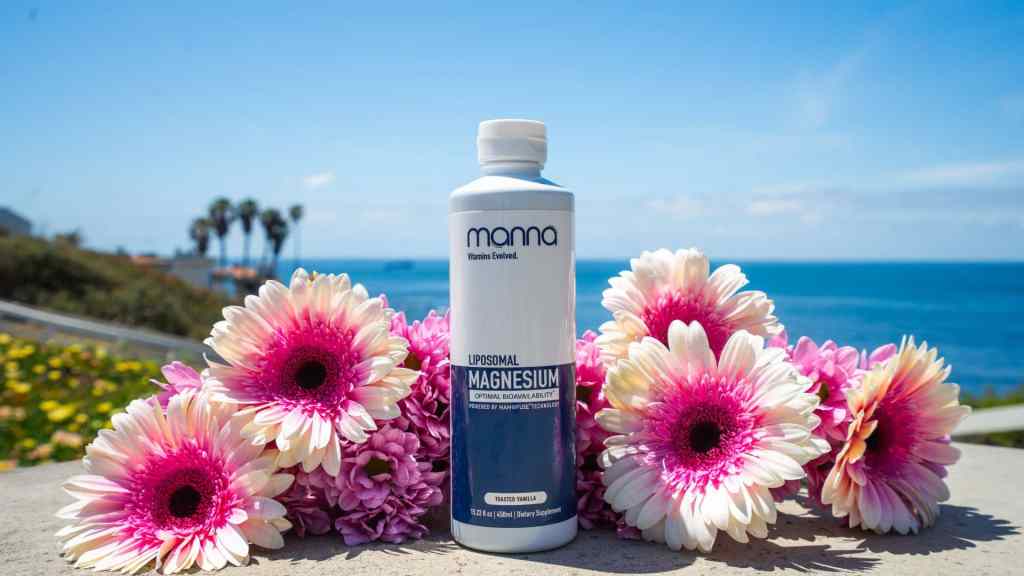 A bottle of Mama Liposomal Magnesium supplement stands on a ledge surrounded by pink and white flowers, with the ocean and palm trees visible in the background.