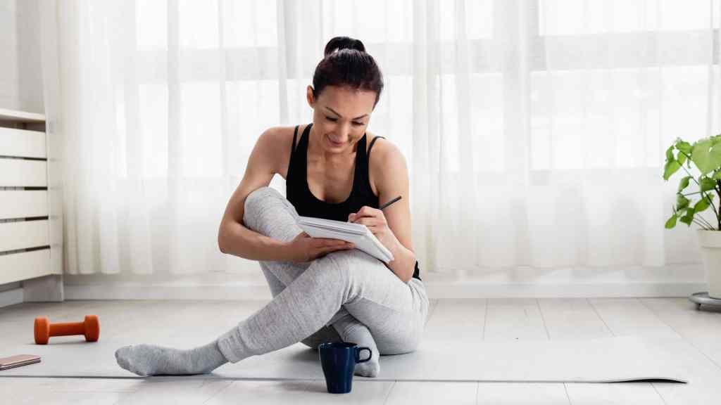 A woman sits on a yoga mat, writing in a notebook with a pen. She wears a black tank top and gray pants. Nearby are a dumbbell and a cup, with a plant in the background.