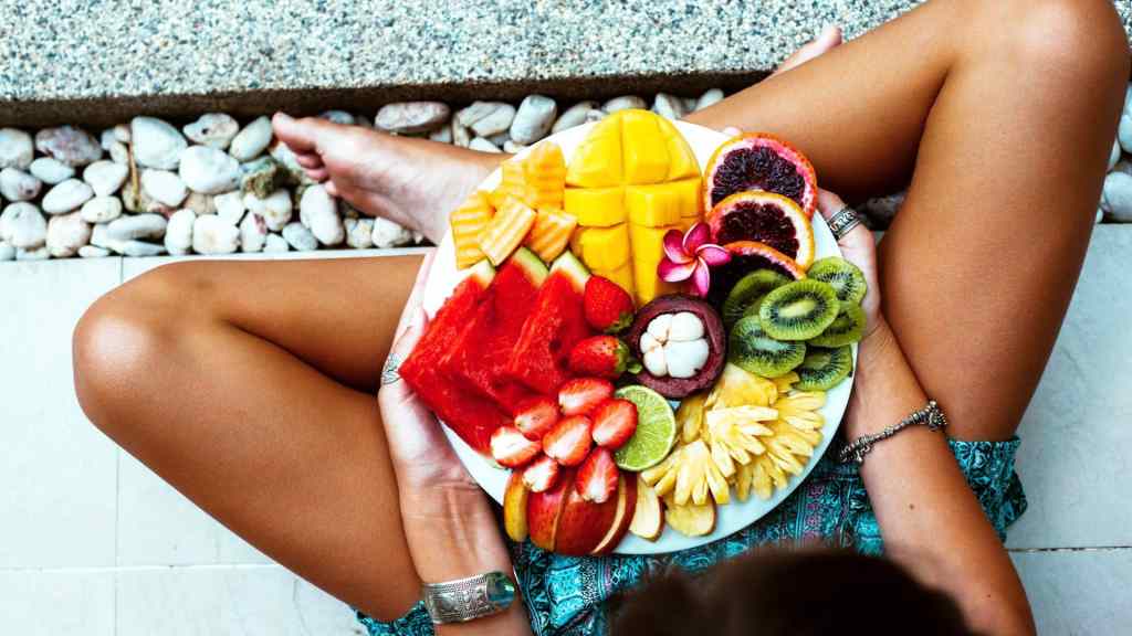Person holding a plate with assorted fruits, including watermelon, mango, kiwi, strawberry, and apple, while seated cross-legged on a stone surface.