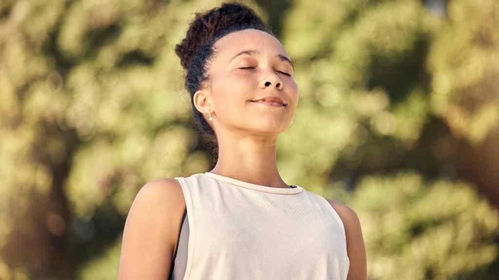 A person with closed eyes stands outdoors in a sunlit area, appearing serene. They wear a sleeveless top, and the background is filled with greenery.