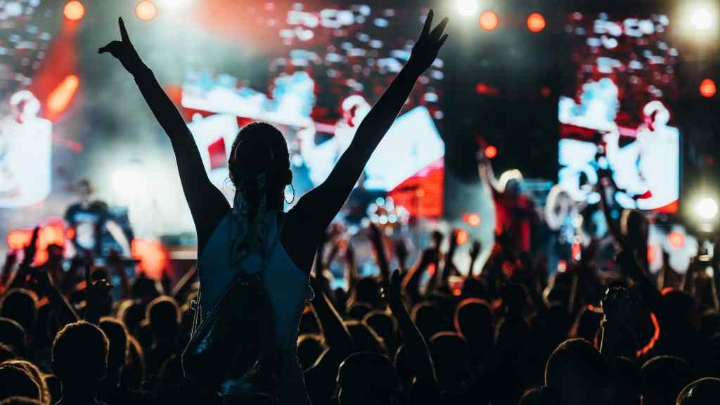A crowd enjoys a live concert, with a silhouette of a person raising arms in the foreground. Bright stage lights illuminate the background.