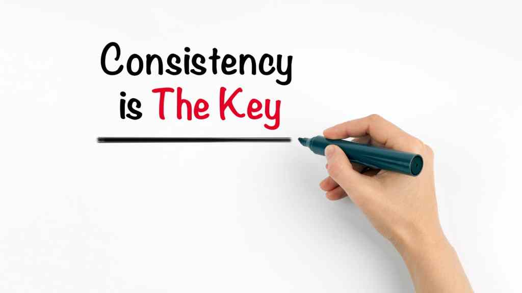 A hand holds a black marker, writing "Consistency is The Key" on a whiteboard, with "The Key" in red and underlined.