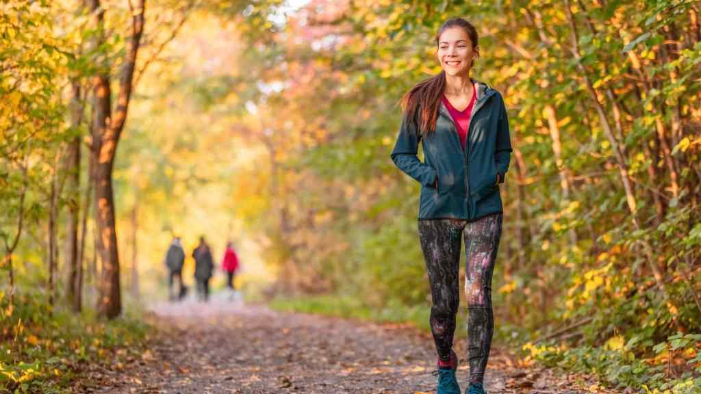 A woman in athletic wear walks on a leaf-covered path in a forest during autumn. Trees with colorful foliage surround her. Three people are blurred in the background.