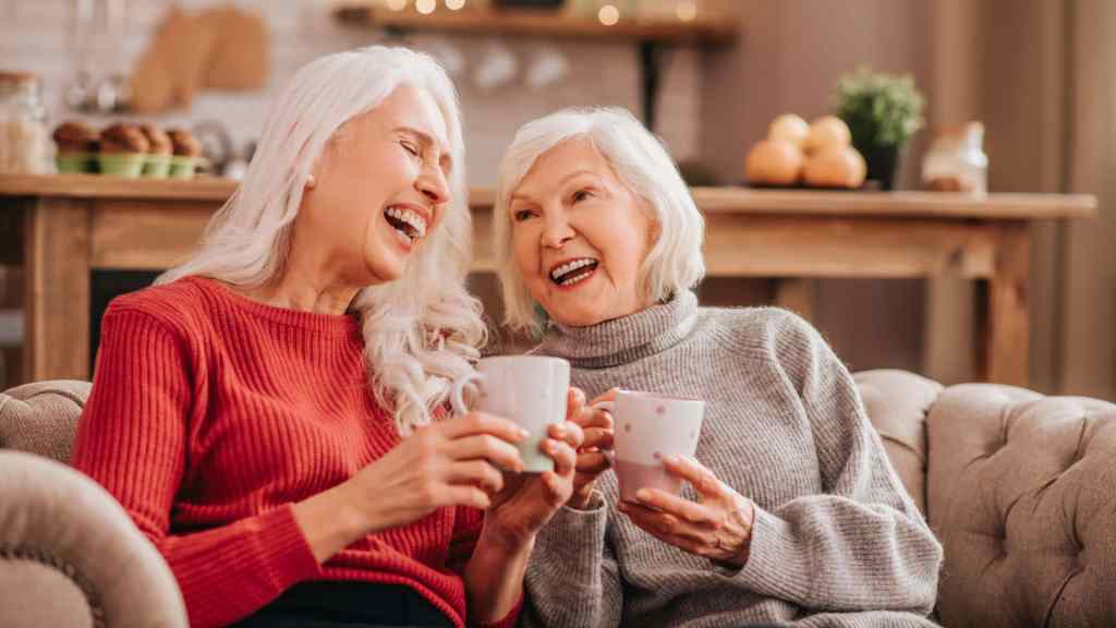 Two older women with gray hair sit on a couch, smiling and laughing together while holding mugs in a cozy, warmly lit living room.