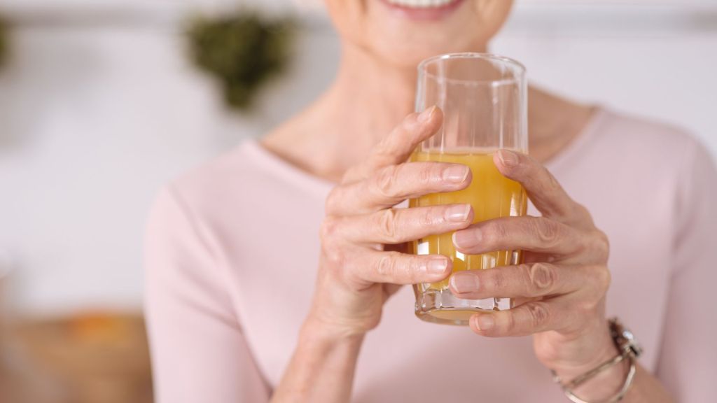 A person wearing a light pink top is holding a glass of orange juice with both hands. Only the lower half of their face and hands are visible.