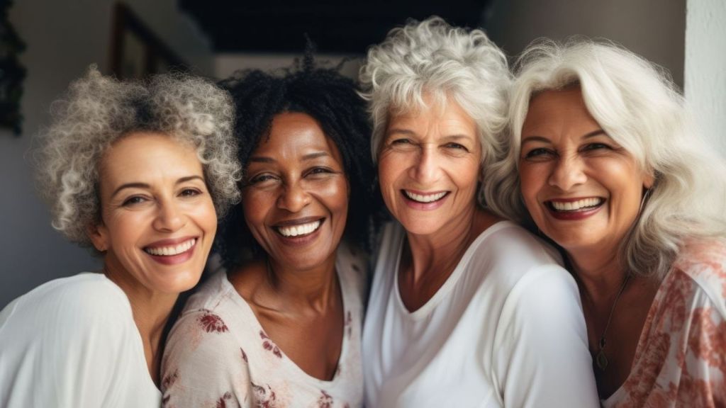 Four older women with gray and white hair stand close together indoors, smiling at the camera.