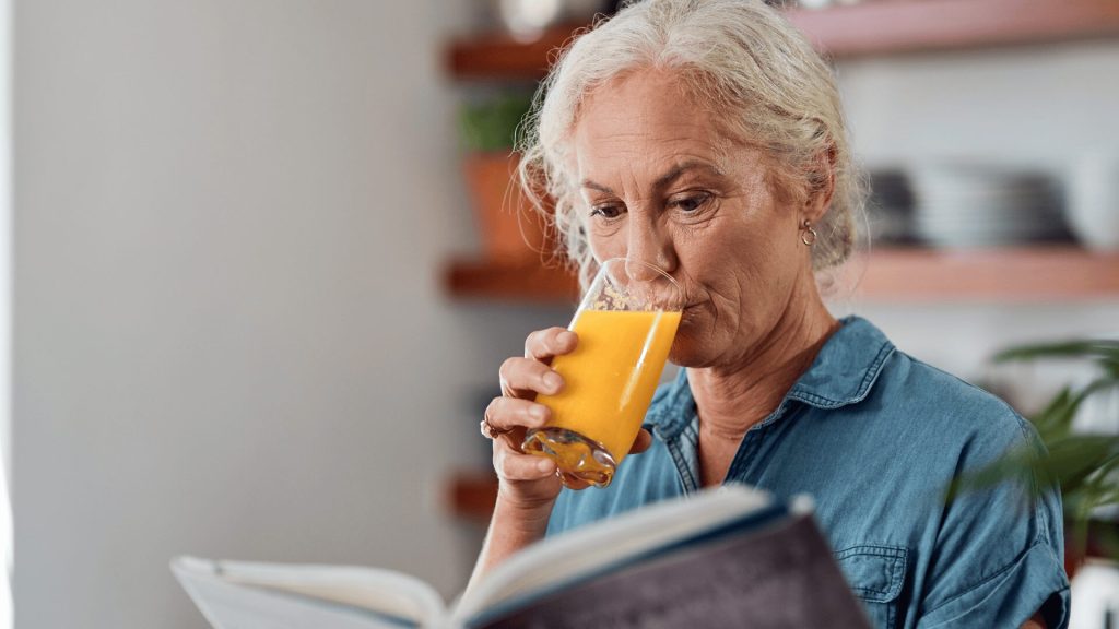 Older woman with gray hair wearing a blue shirt drinks orange juice while reading a book indoors.