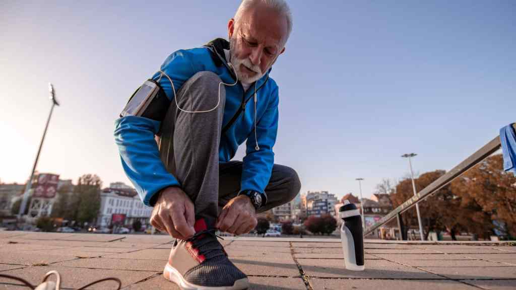 Older man in athletic wear ties his shoelaces outdoors, with a water bottle nearby. He stands on a paved area with a cityscape and trees in the background.