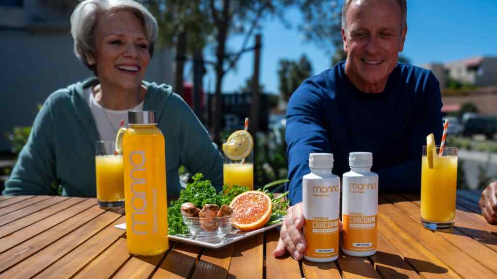 Two older adults sit at an outdoor table with juice, a grapefruit, greens, and bottles labeled "manna" and "curcumin" dietary supplement.