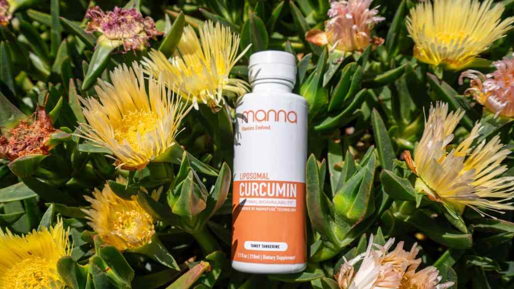 A bottle of Manna Liposomal Curcumin dietary supplement is placed among yellow and pink blooming flowers and green foliage.