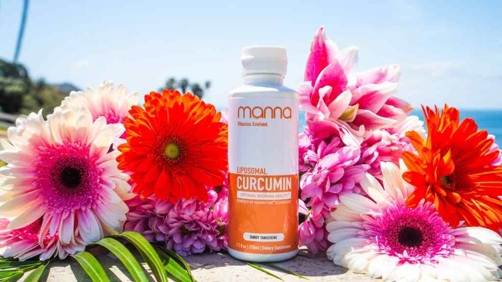 A bottle of Mama Liposomal Curcumin supplement is surrounded by vibrant pink, orange, and red flowers with a blue sky and ocean in the background.