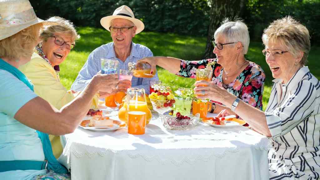 Five older adults sit around a table outdoors, smiling and toasting drinks, with fruit and juice on the table, on a sunny day.