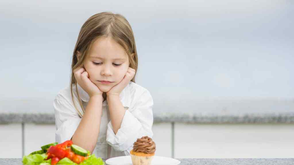 A child looks at a plate with a cupcake and a bowl of salad, appearing thoughtful.