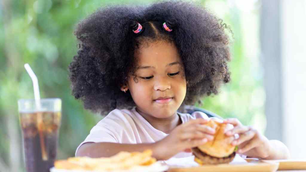 Child eating a burger at a table with fries and a soda in an outdoor setting.