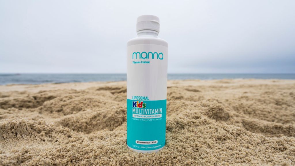 A bottle of Manna Liposomal Kids Multivitamin stands upright on a sandy beach with a cloudy sky and ocean in the background.