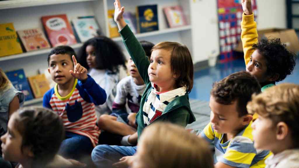 Children seated on a classroom floor raise their hands, surrounded by books on a shelf.