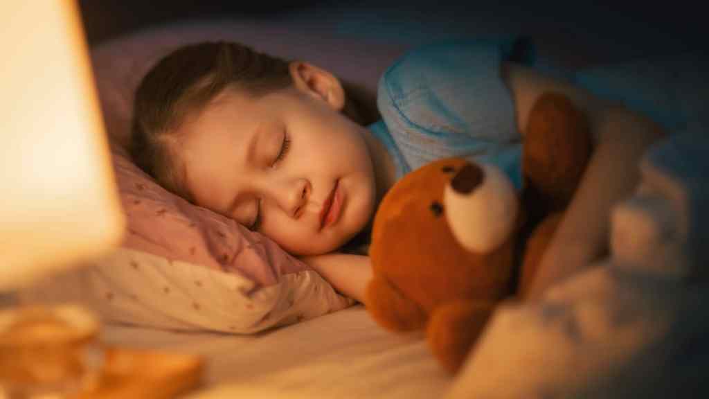 Child sleeping peacefully in bed, holding a teddy bear. A warm light illuminates the scene.