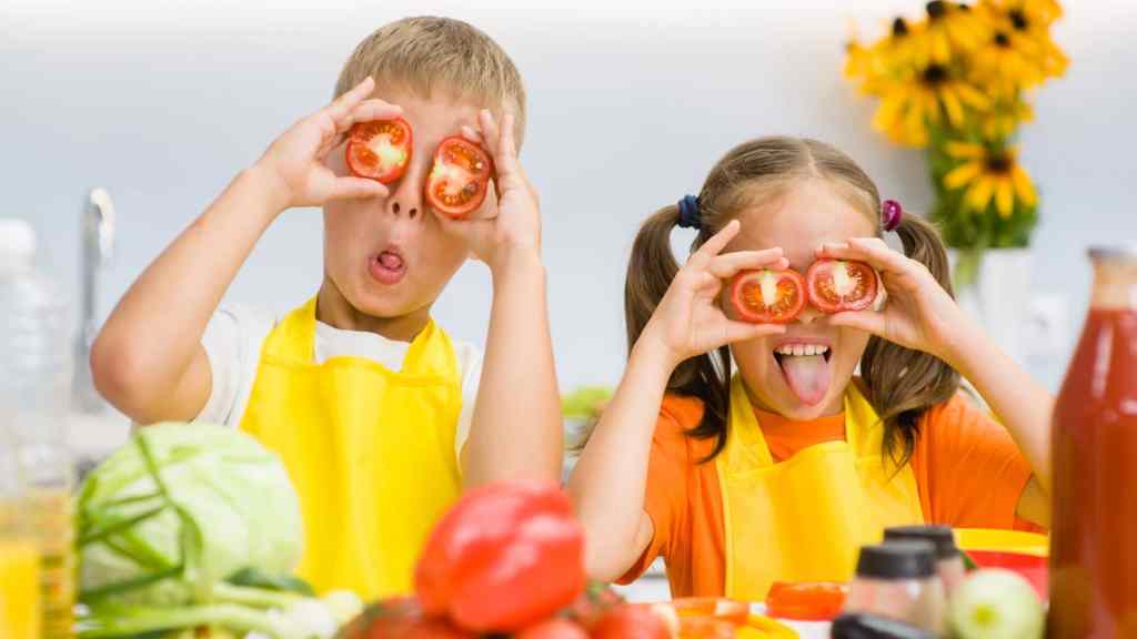 Two children in yellow aprons hold tomato slices over their eyes, making playful faces. A variety of vegetables and flowers are visible in the foreground and background.