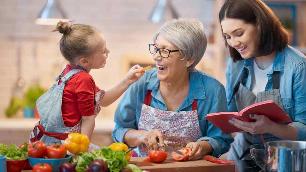 A girl feeds a slice of vegetable to an older woman while another woman smiles, holding a cookbook. They are in a kitchen with various vegetables on the counter.