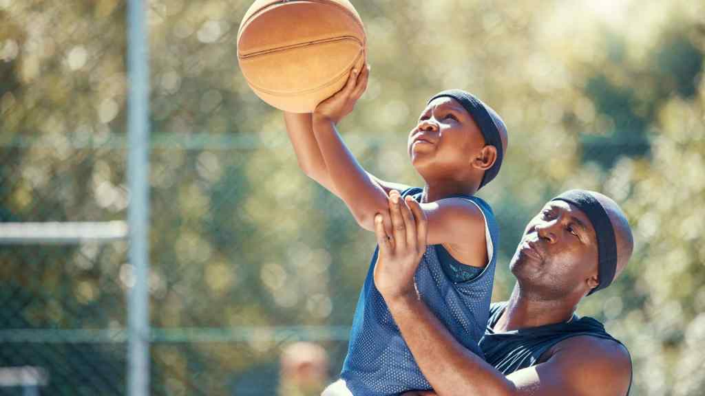 A man assists a young boy in shooting a basketball outdoors. Both wear headbands and athletic clothing.