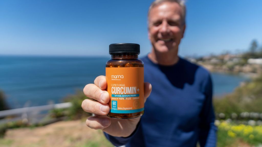 A person outdoors holds a bottle of mama Universal Curcumin+ supplement in front of the camera with the ocean and coastal scenery in the background.