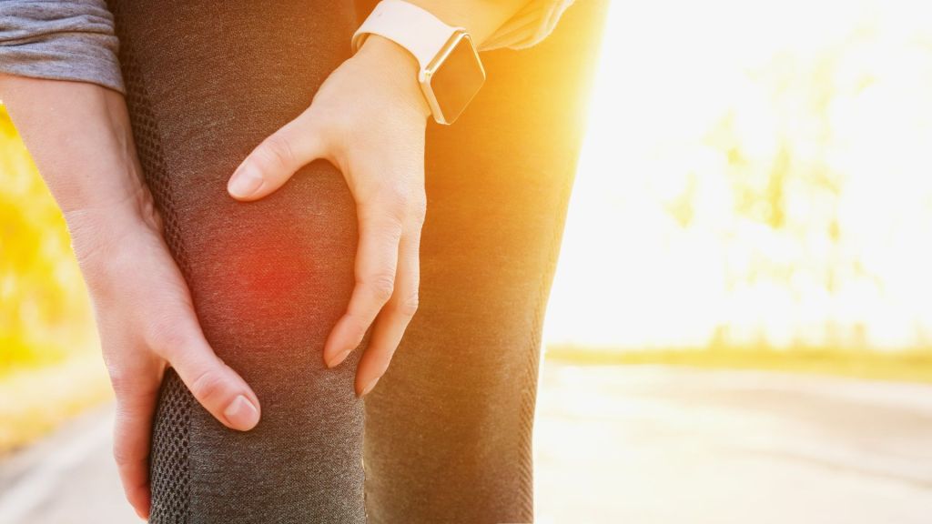A person wearing a smartwatch holds their knee in pain, illuminated by warm sunlight.