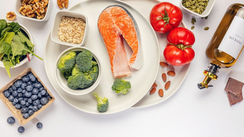 A variety of healthy foods, including salmon, tomatoes, broccoli, blueberries, spinach, nuts, oatmeal, olive oil, and seeds, are arranged on a white surface.