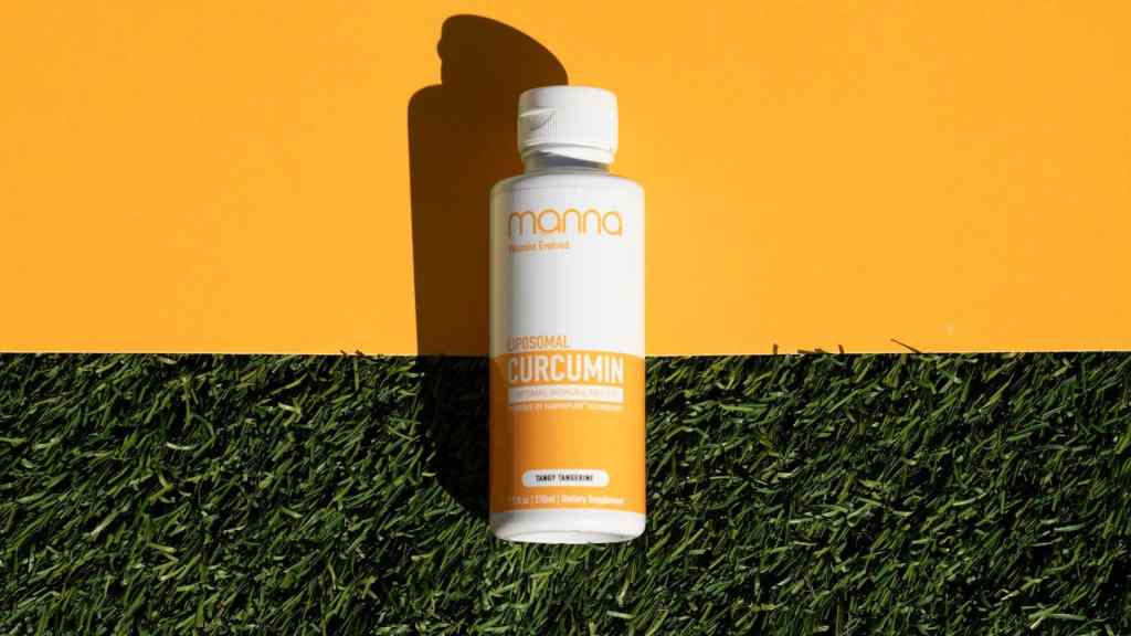 A white bottle labeled "Liposomal Curcumin" lies on a background split between yellow at the top and green grass at the bottom, casting a shadow.