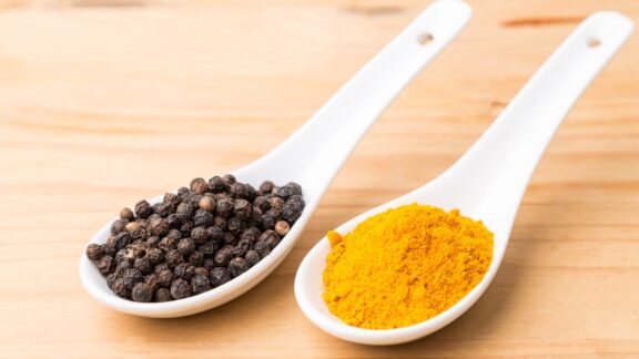 Two white ceramic spoons on a wooden surface: one contains black peppercorns, the other holds yellow turmeric powder.