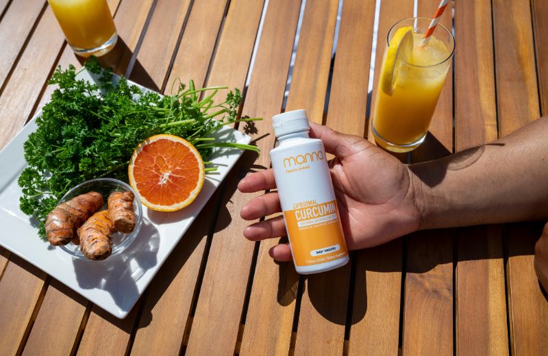 A hand holding a bottle of curcumin supplement next to a plate with turmeric roots, an orange, and parsley, with two glasses of juice on a wooden table.
