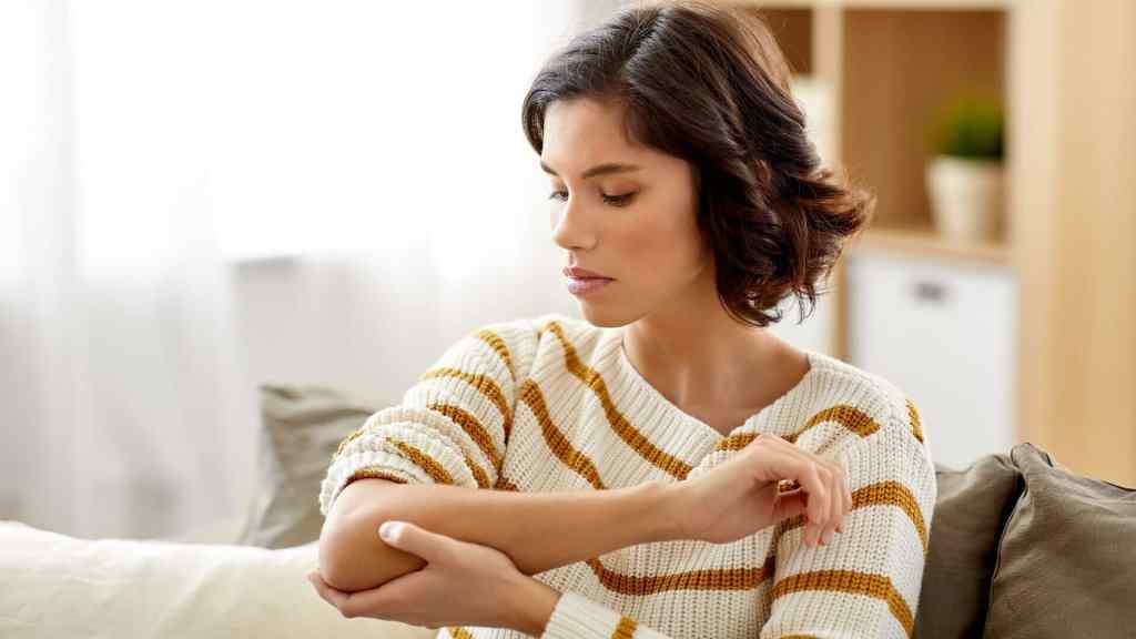 A woman in a striped sweater sits on a couch, examining her elbow with a concerned expression.
