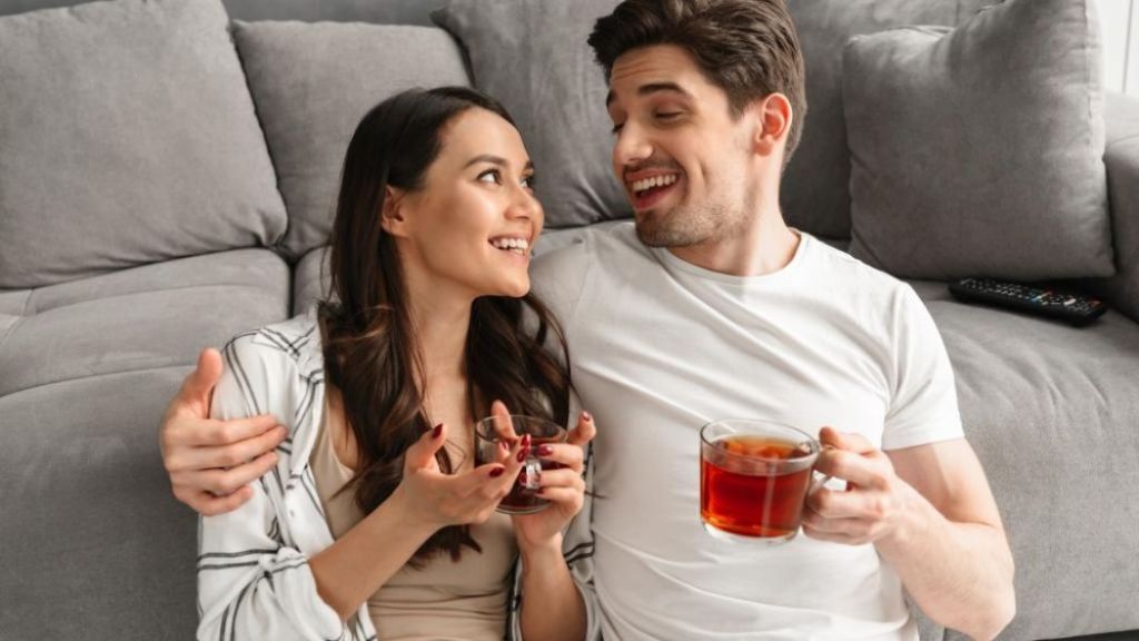 A couple sitting on a sofa, smiling and having a conversation with drinks in their hands.