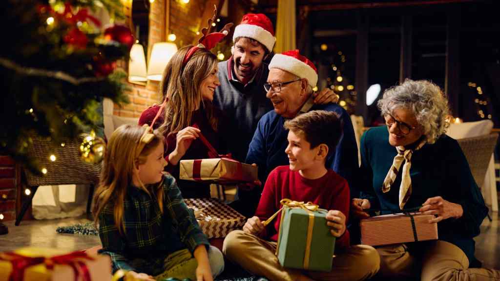 A family sits together near a Christmas tree, exchanging gifts and wearing festive hats.