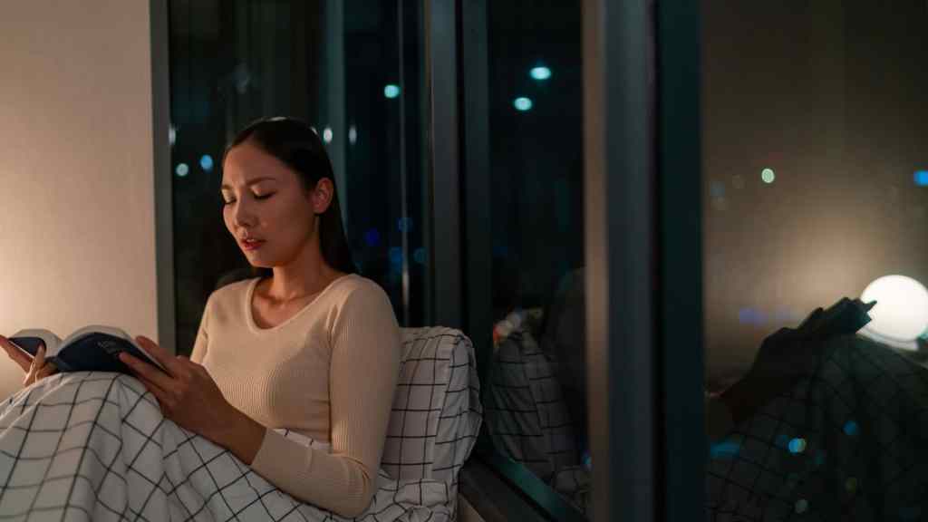A woman in a beige sweater sits by a window, reading a book at night. A lamp provides soft light, and city lights are visible outside.