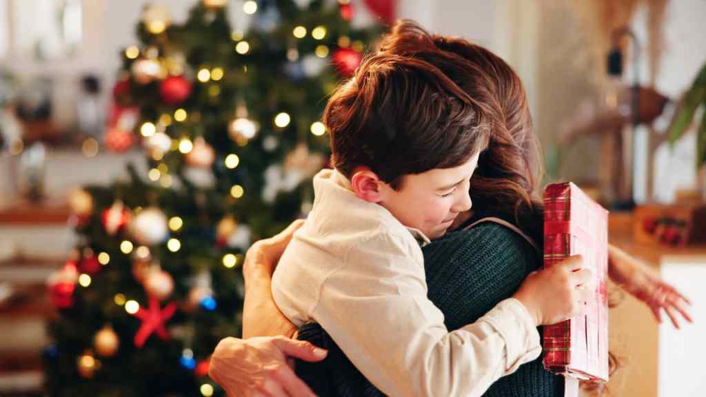 A child and an adult hugging in front of a decorated Christmas tree; the child holds a wrapped gift.