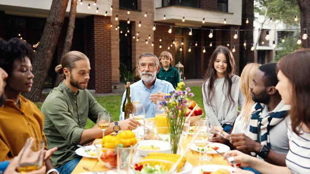 A diverse group of people enjoy a meal outdoors with drinks, string lights, and a variety of dishes on the table. A brick building is in the background.