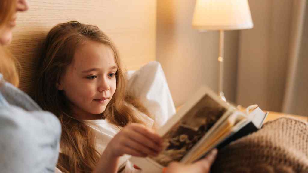 A girl lying in bed reads a book, with a lamp lit in the background.