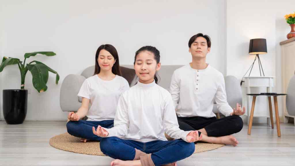 Three people sitting cross-legged and meditating in a living room. Two adults are behind a child, all with eyes closed.