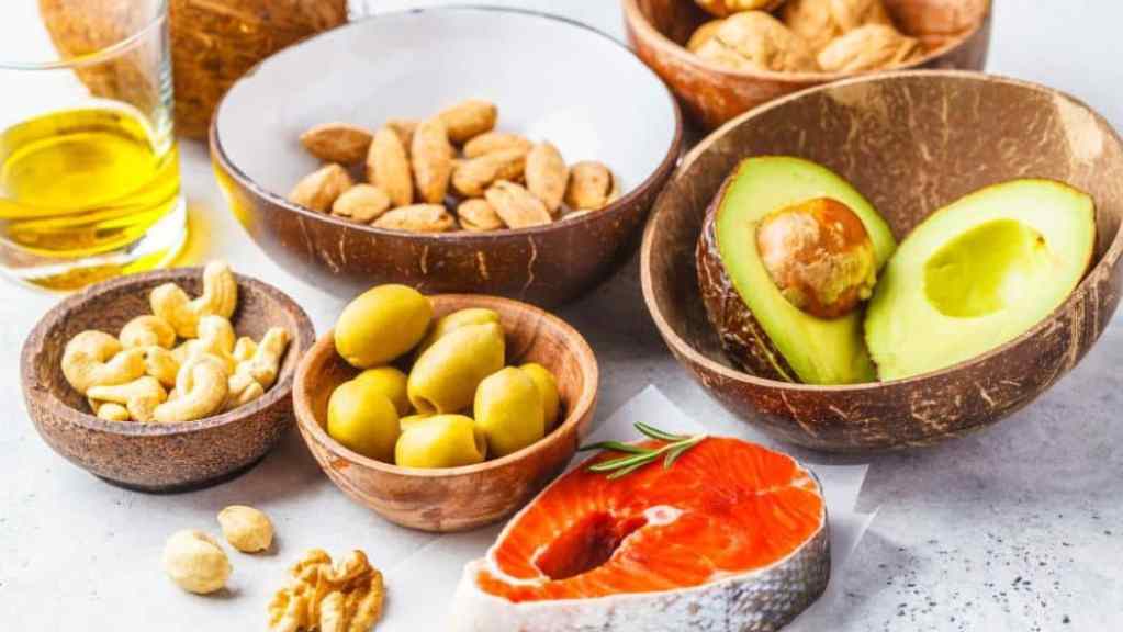 A variety of healthy foods including nuts, olives, avocado, and salmon, arranged in bowls on a light surface.