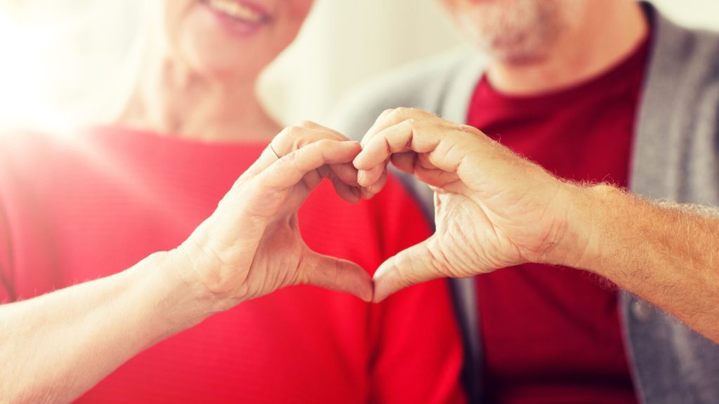 Two people sitting close together form a heart shape with their hands. Both are wearing red tops.