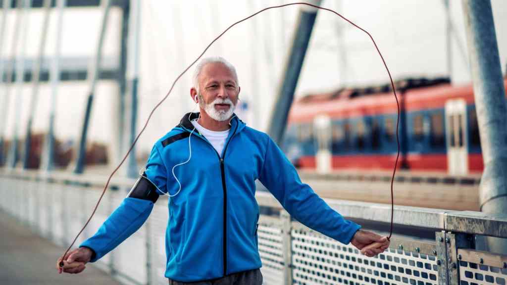 Elderly man exercising by jumping rope on a bridge, wearing a blue jacket and headphones, with a train passing in the background.
