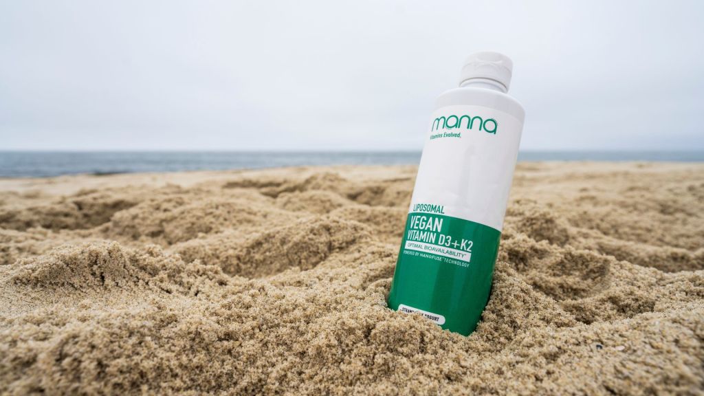 A bottle of vegan vitamin D3+K2 is partially buried in sand on a beach, with the ocean in the background.