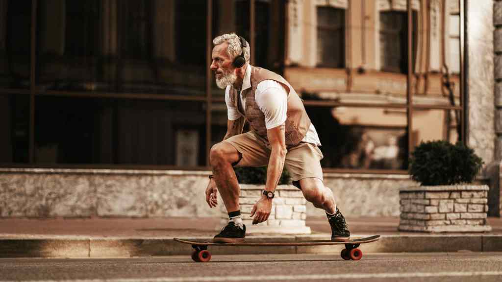 Older man with a beard, wearing headphones, a vest, and shorts, skateboards on a street near a building.