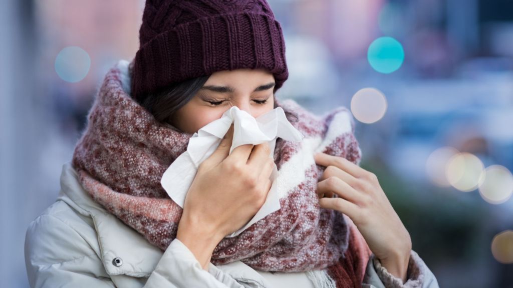 Person wearing a purple beanie and scarf is blowing their nose with a tissue.