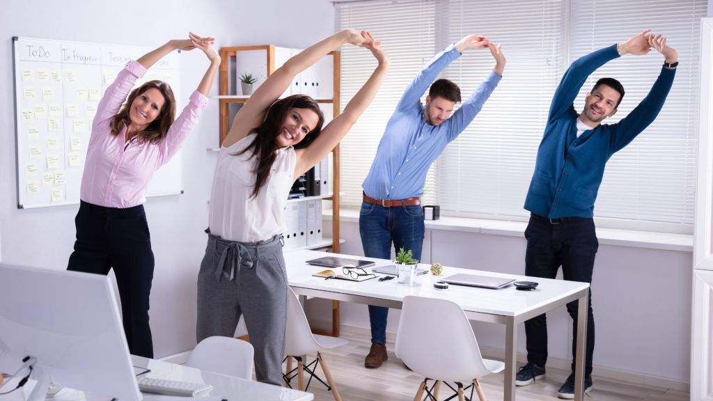 Four people in business attire are standing and stretching with their arms raised in an office setting, next to desks with computers and other office equipment.