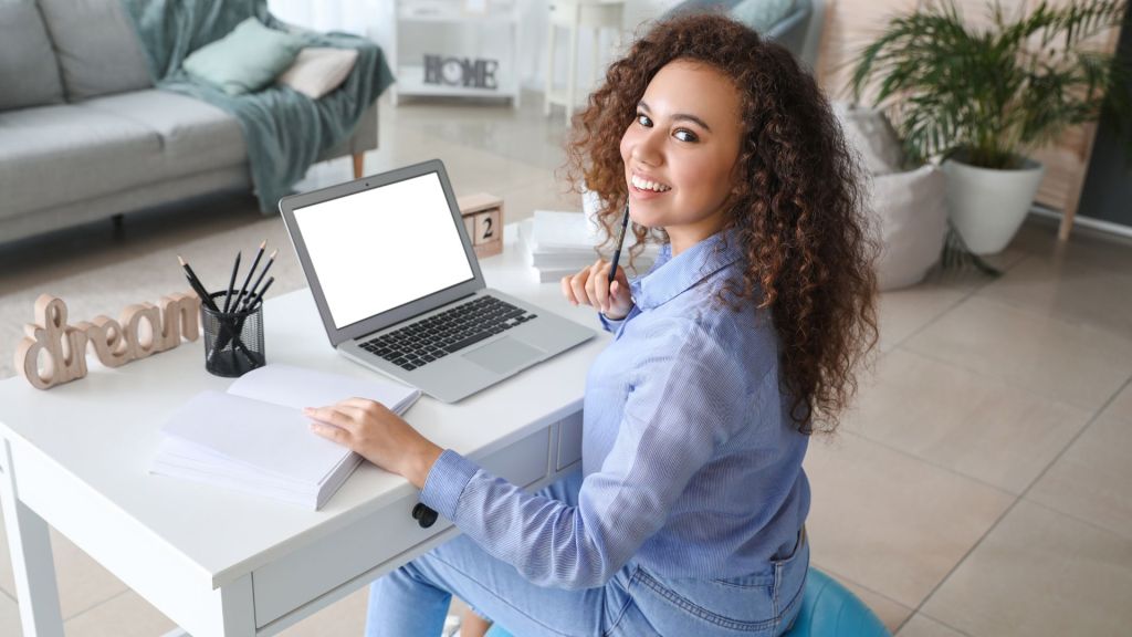 A young woman with curly hair smiling at the camera while seated at a desk with a laptop, notebook, and a 'dream' sign in a cozy living room.