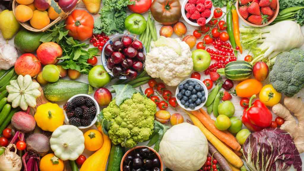Assorted fresh fruits and vegetables, including tomatoes, berries, squash, cabbage, apples, carrots, broccoli, and peppers, arranged closely together on a flat surface.